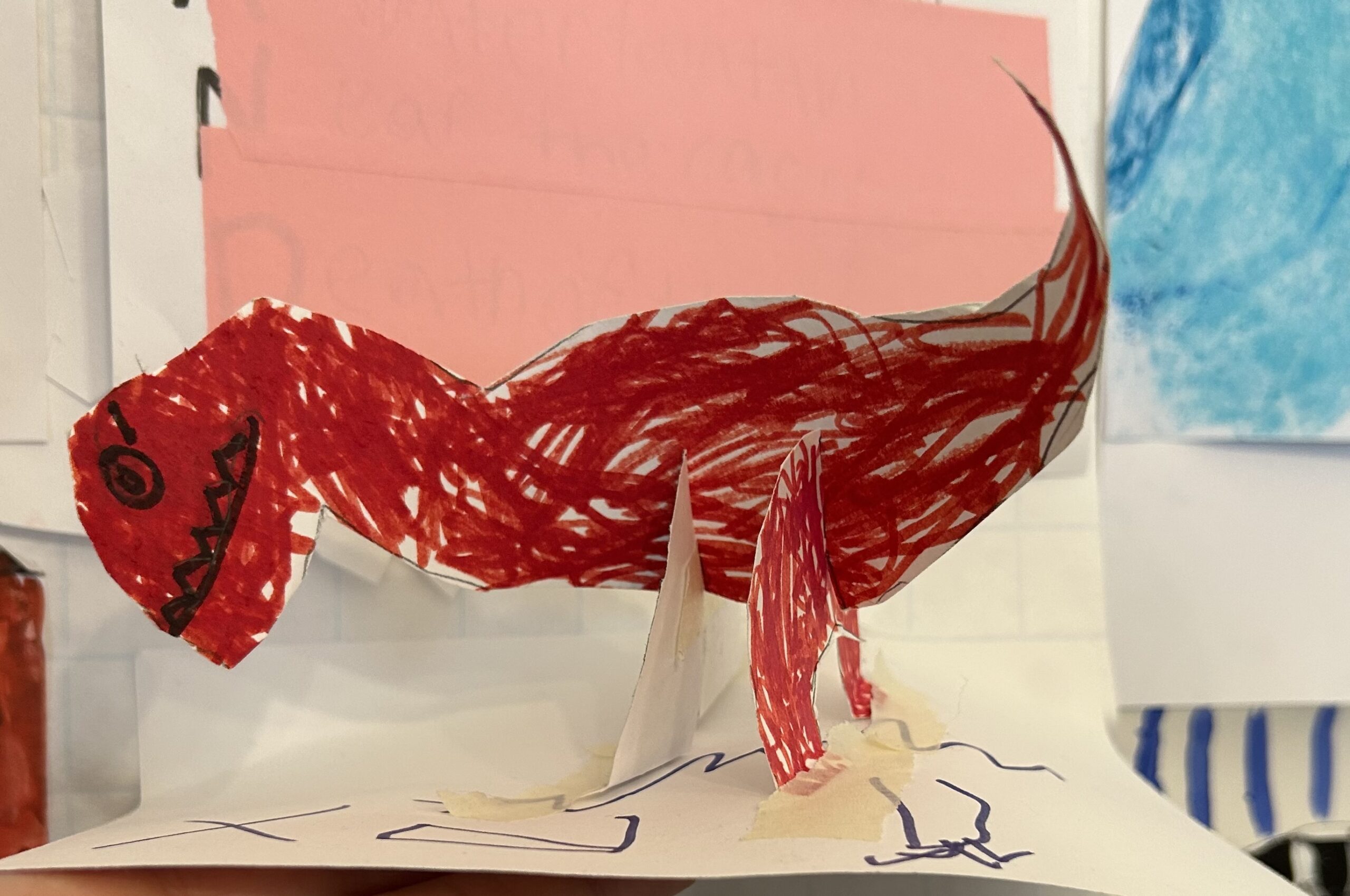 a crayon drawn and cut out from paper red tyrannosaurus rex with paper legs taped onto a flat service to appear standing, made by a child